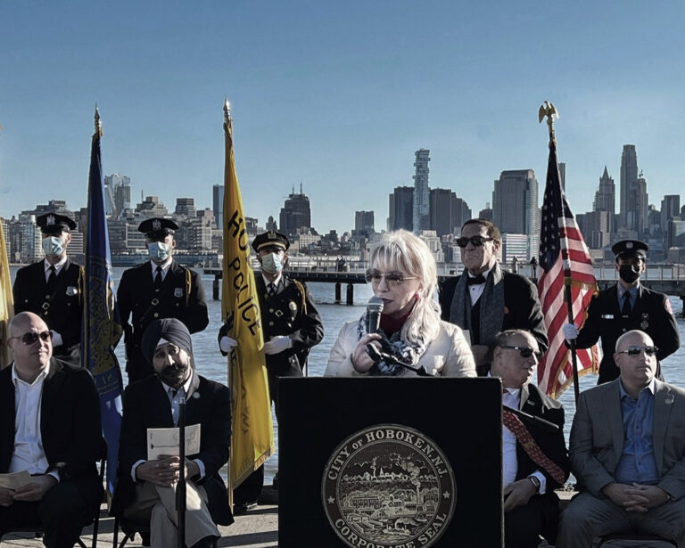 Sculptor Carolyn D. Palmer speaking at a podium during an outdoor unveiling event, with uniformed attendees and flags behind her.