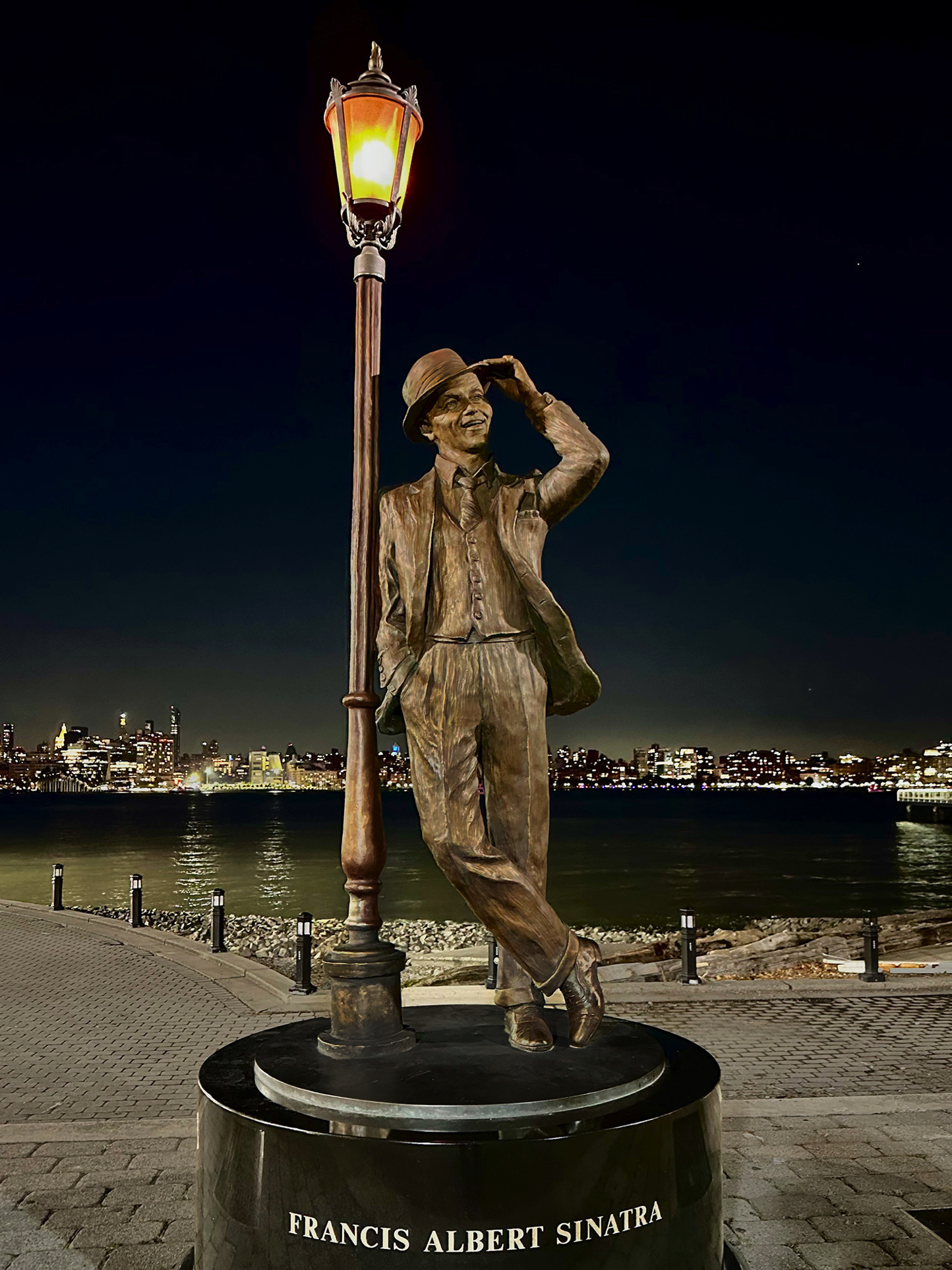 Full-length view of the bronze Frank Sinatra statue beneath a lit lamppost at night, set along a waterfront promenade.