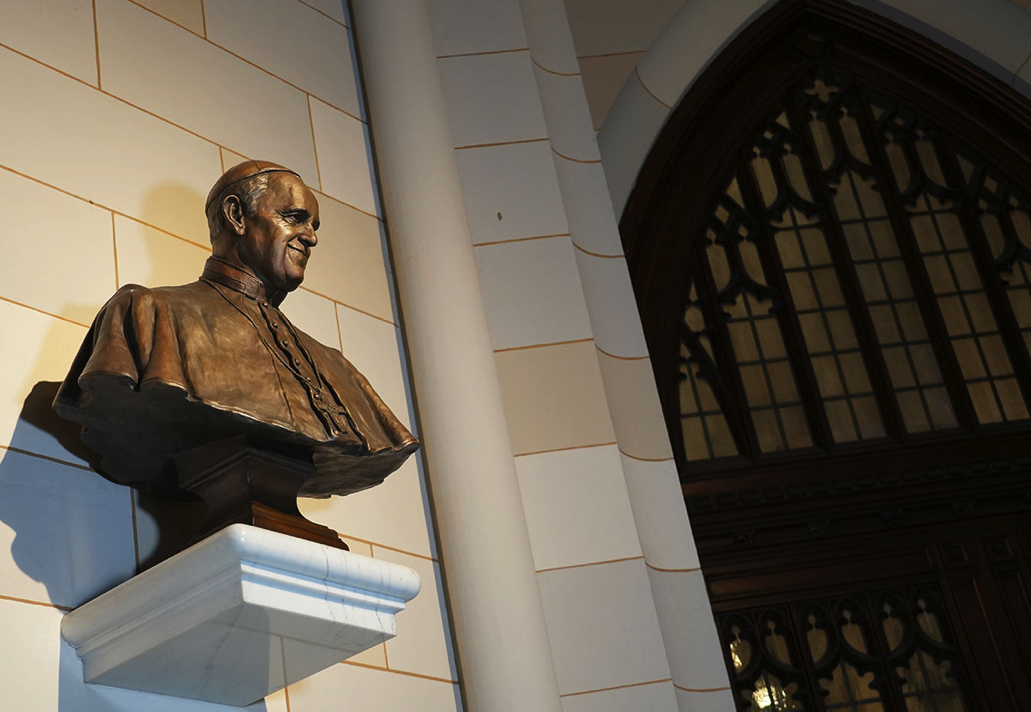 Bronze bust of Pope Francis mounted on an exterior wall near an arched entrance, viewed from below.