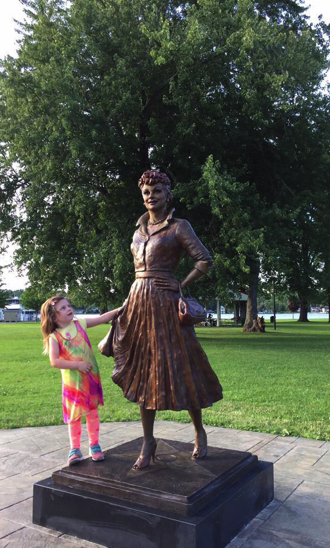 Child standing beside the bronze Lucille Ball statue, reaching toward the sculpture in a park setting.
