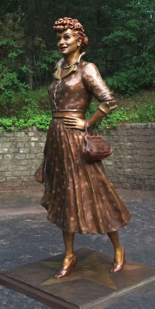 Full-length bronze statue of Lucille Ball standing with hands on hips, wearing a dress and heels.