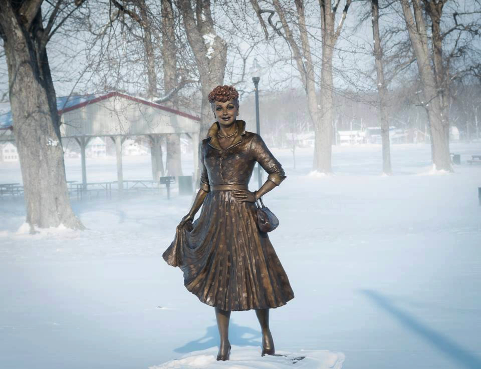 Bronze Lucille Ball statue standing outdoors in a snowy park, surrounded by bare trees.