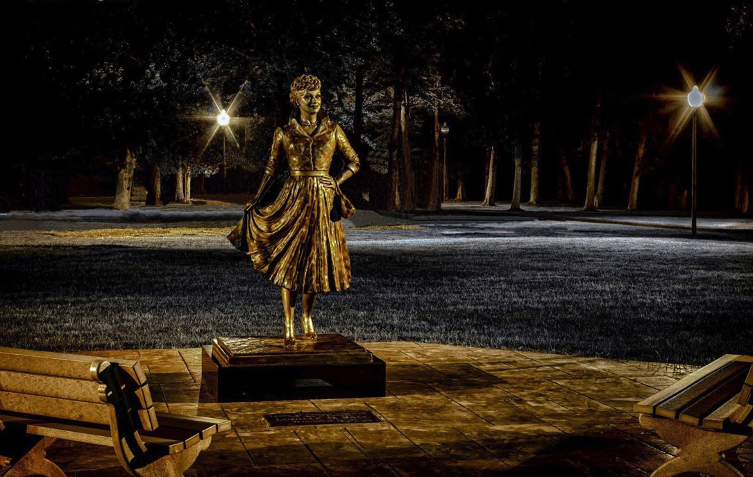 Bronze statue of Lucille Ball standing on a pedestal in a park at night, illuminated by nearby lampposts.