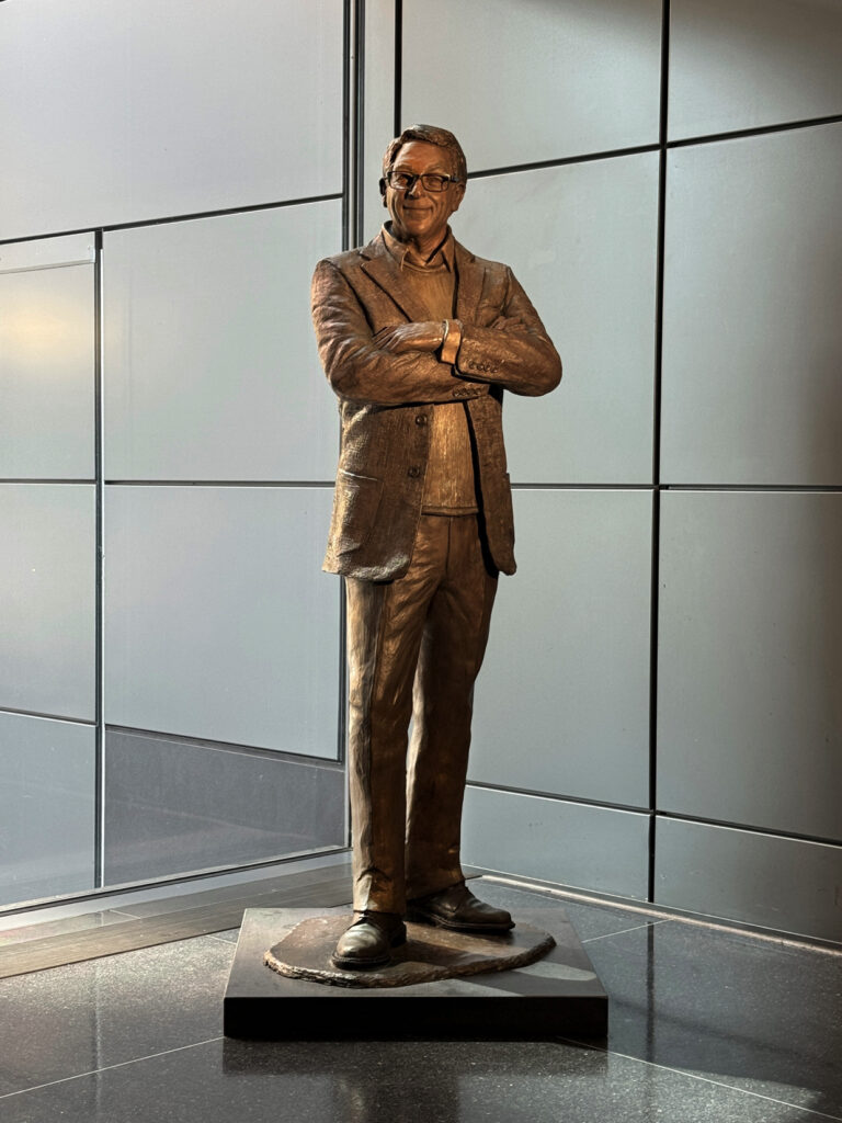 Full-length bronze statue of Stuart Weitzman standing with arms crossed on a pedestal in a modern interior space.