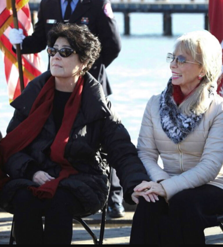 Carolyn Palmer and Tina Sinatra seated and holding hands during an outdoor sculpture unveiling ceremony.
