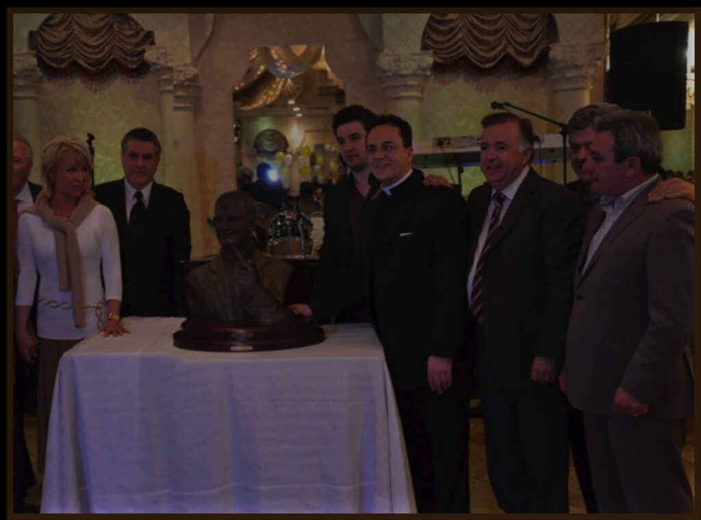 Group of formally dressed attendees standing around a table displaying a bronze bust at an indoor unveiling event.