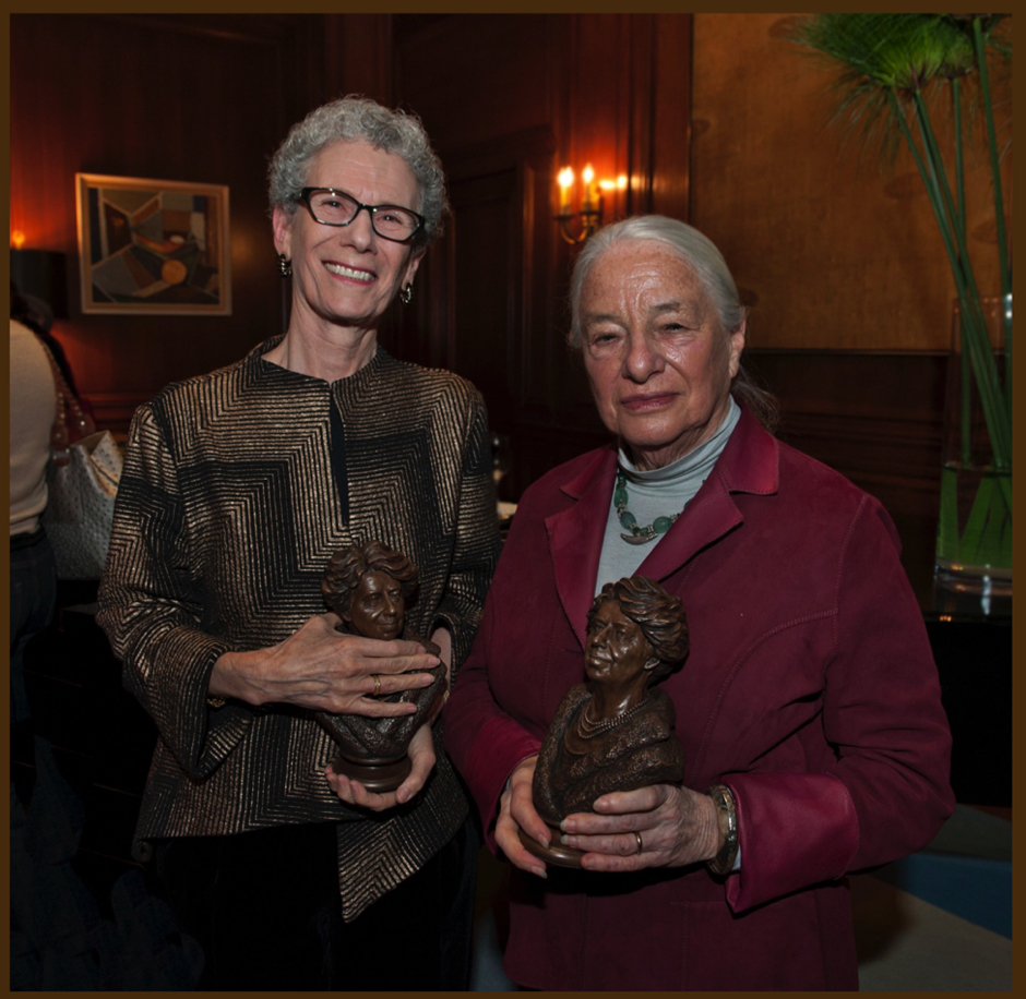 Two women standing indoors holding small bronze bust trophies during a presentation event.