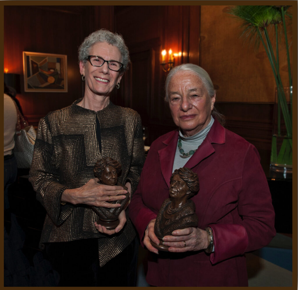 Two women standing indoors holding a small bronze bust during a presentation event.