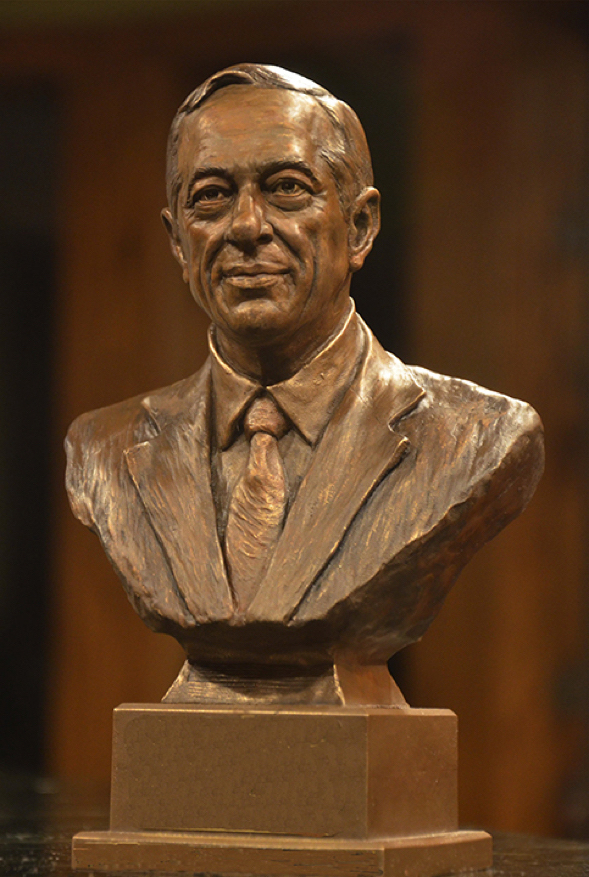 Front view of a bronze bust depicting Mario Cuomo, mounted on a pedestal against a dark background.