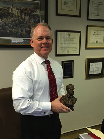 Man standing in an office holding a tabletop bronze bust of Sir William Osler, with framed certificates on the wall behind him.