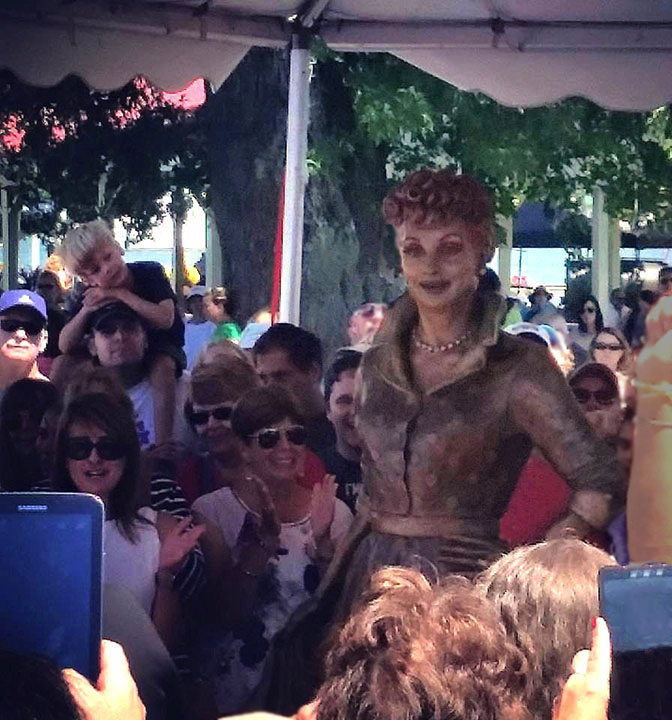 Bronze Lucille Ball statue surrounded by a crowd during an outdoor unveiling ceremony.