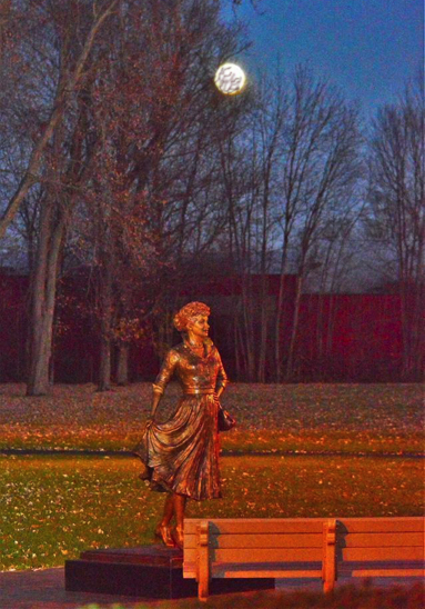 Bronze Lucille Ball statue displayed in a park during early evening, with autumn trees and benches nearby.
