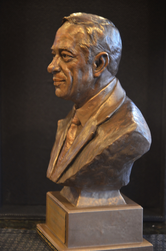 Profile view of a bronze bust of Mario Cuomo, showing the sculpted hair, suit collar, and tie.