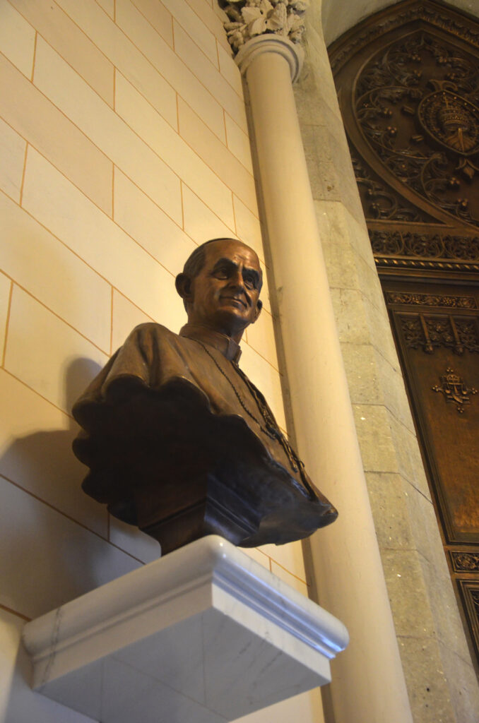 Bronze Pope Paul bust displayed on a wall pedestal inside St. Patrick’s Cathedral, viewed from a lower angle near an ornate doorway.