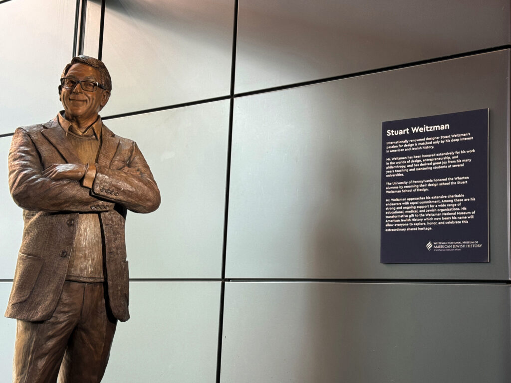 Bronze statue of Stuart Weitzman displayed beside a wall-mounted informational plaque in a contemporary gallery setting.