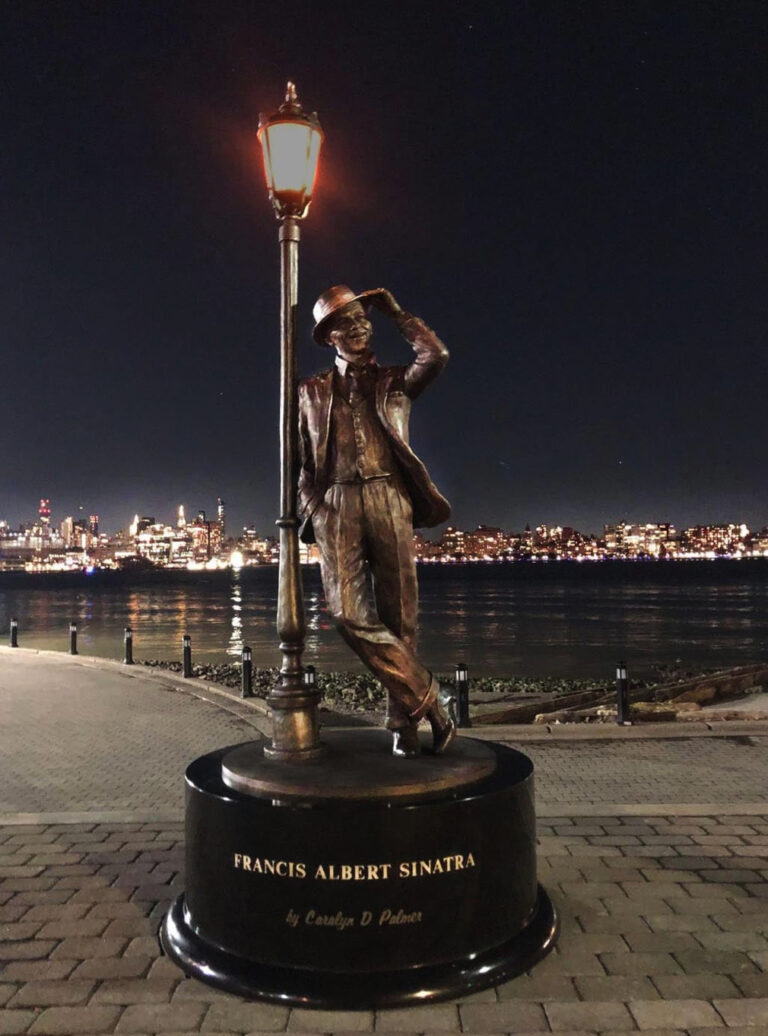 Vertical nighttime view of the bronze Frank Sinatra statue and lamppost, with illuminated city skyline behind.