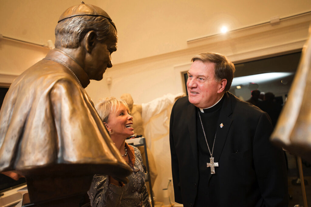 Sculptor Carolyn D. Palmer speaking with a Catholic cardinal beside a bronze papal bust.