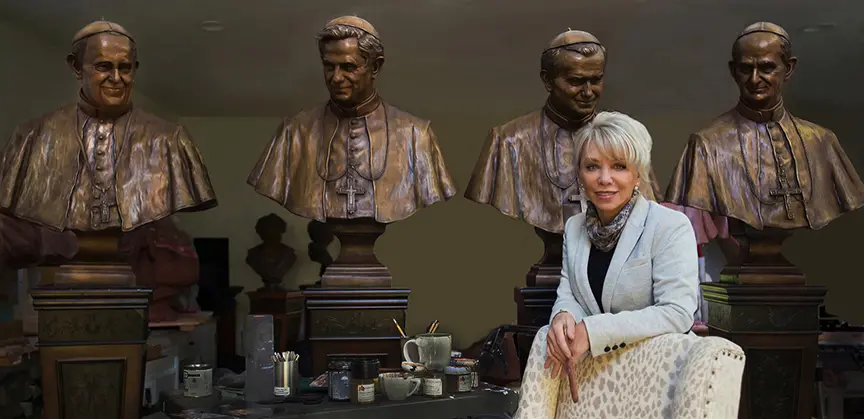 Sculptor Carolyn D. Palmer seated in her studio surrounded by multiple bronze busts arranged on worktables behind her.